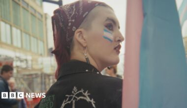 Mason is at a rally and looking over his shoulder. He is wearing make-up and a dark-red bandana. He has a blue, pink and white transgender Pride flag painted on his face.