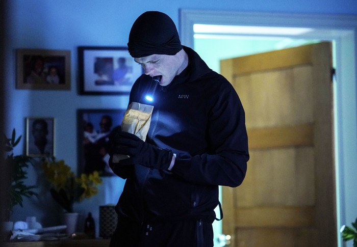 Oscar dressed in a black hat and outfit holding an envelope and torch in EastEnders