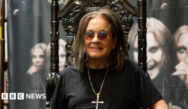Ozzy Osbourne - a man with long dark hair, sunglasses, a necklace with a crucifix attached, and a black shirt, sits on a black gothic-style throne and smiles. Behind him is a large banner featuring multiple images of him.
