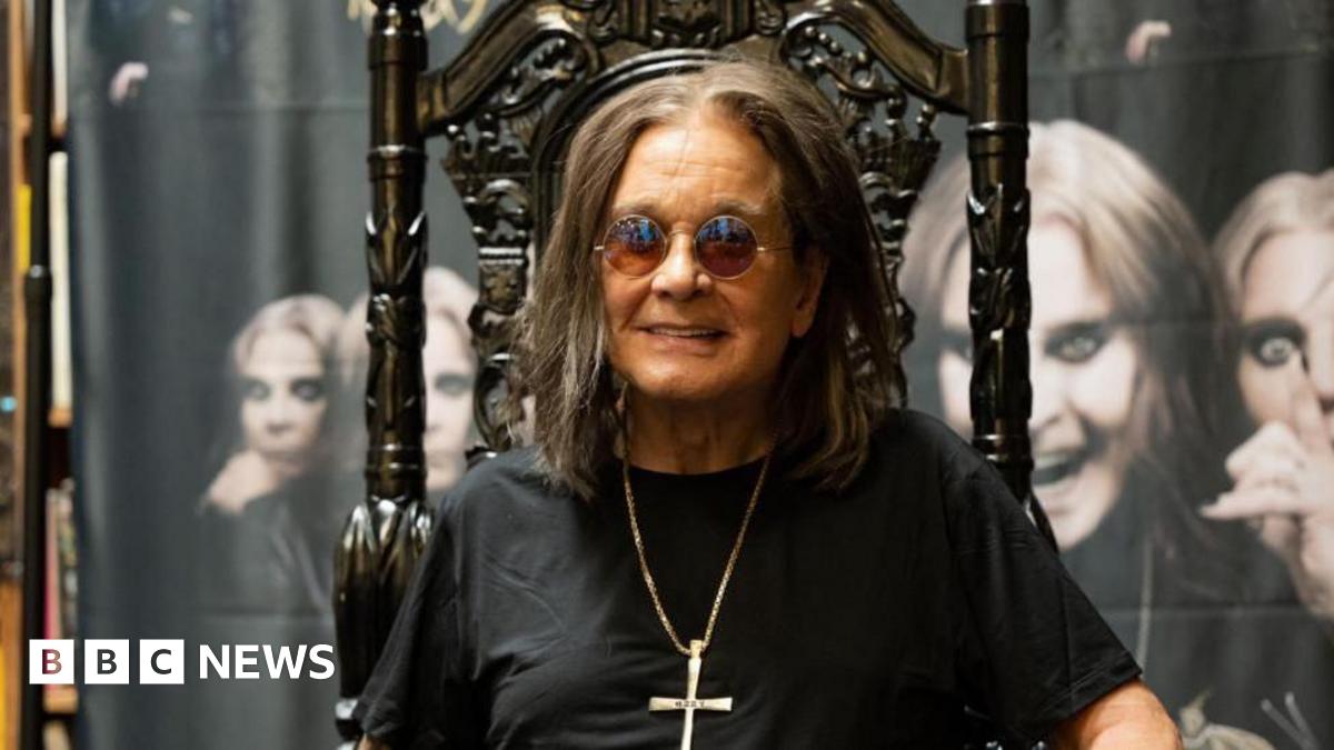 Ozzy Osbourne - a man with long dark hair, sunglasses, a necklace with a crucifix attached, and a black shirt, sits on a black gothic-style throne and smiles. Behind him is a large banner featuring multiple images of him.