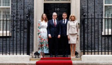 Four people stand on a red carpet in front of the iconic black door of 10 Downing Street, a historic brick building with large windows, in London.