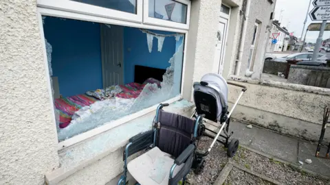 PA Media A black wheelchair and white pram sit outside a smashed ground floor window. On the other side of the smashed glass is a blue painted bedroom, with a black bedframe and pink bed sheets. 