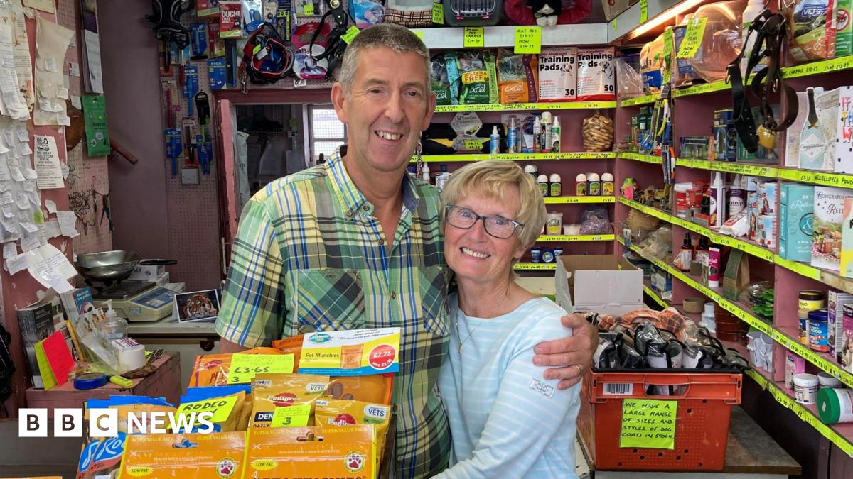Terry has short hair and is wearing a green checked shirt. He is standing with his arm around Orla who has short blonde hair and is wearing glasses and a blue and white striped top. They are both stood at the counter in the pet shop smiling at the camera.