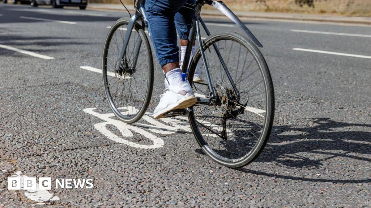 A road surface with a cycle lane marked out by a white bicycle symbol. A dark bike in the cycle land. All that can be seen of the cyclist is their legs and feet. They are wearing jeans and white trainers.