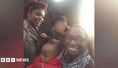 A young baby in a red coat and grey hood is in the middle of a family photo. A woman is holding him and wearing black glasses and a zebra-print scarf while smiling at the camera. A woman behind the baby wears a green coat and red scarf in her hair, while also smiling at the camera. Another figure behind the baby has their face blurred out.