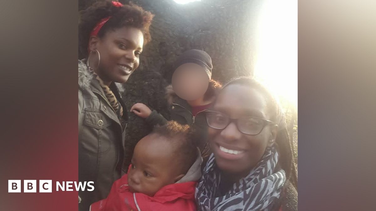 A young baby in a red coat and grey hood is in the middle of a family photo. A woman is holding him and wearing black glasses and a zebra-print scarf while smiling at the camera. A woman behind the baby wears a green coat and red scarf in her hair, while also smiling at the camera. Another figure behind the baby has their face blurred out.