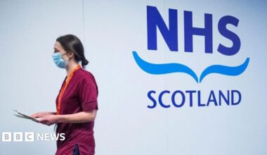 A woman with dark hair, wearing red scrubs and a blue face mask, walks past a white wall with a large NHS Scotland logo on it. She is carrying a clipboard and is side-on to the camera.