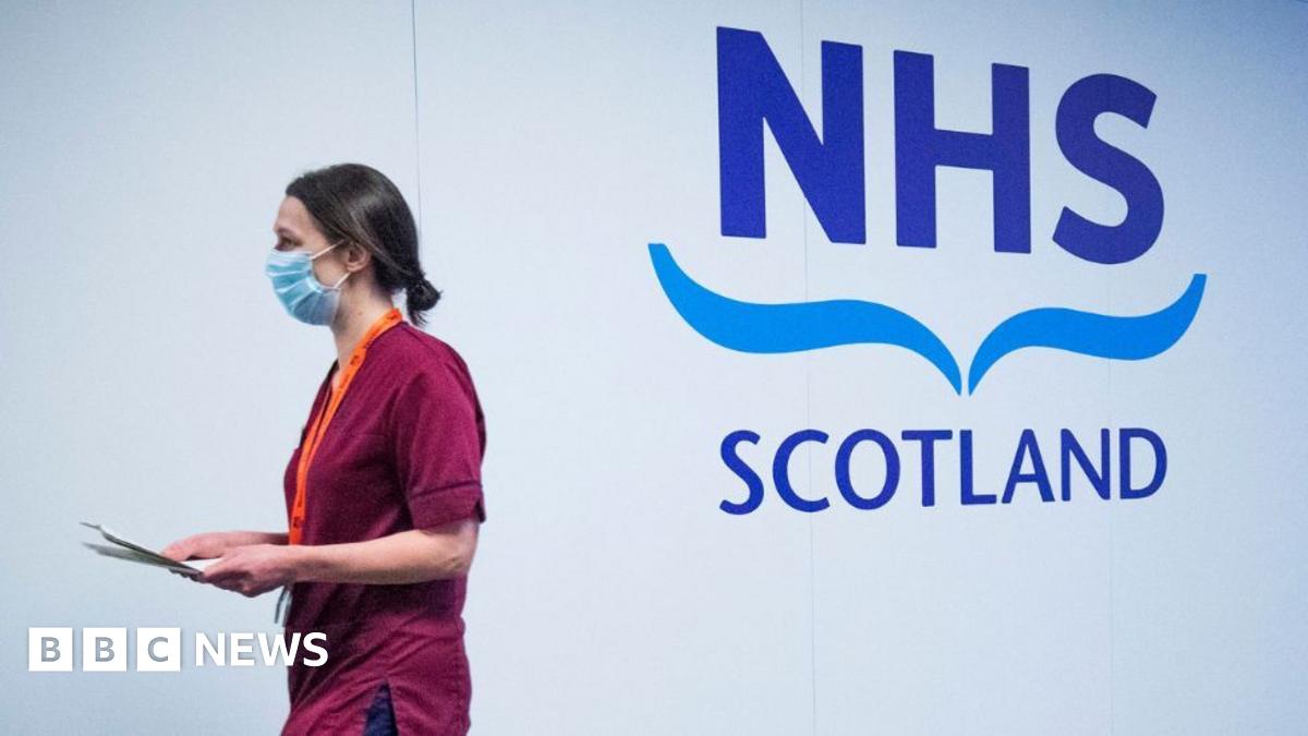 A woman with dark hair, wearing red scrubs and a blue face mask, walks past a white wall with a large NHS Scotland logo on it. She is carrying a clipboard and is side-on to the camera.