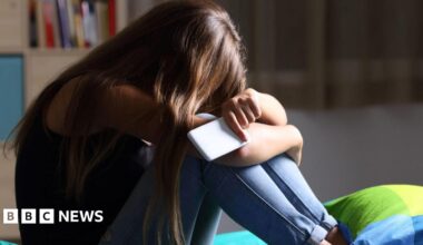 A young woman with long dark hair sits on a bed with her head in her arms. She is holding a white smartphone and is wearing a dark sleeveless top and light blue jeans.