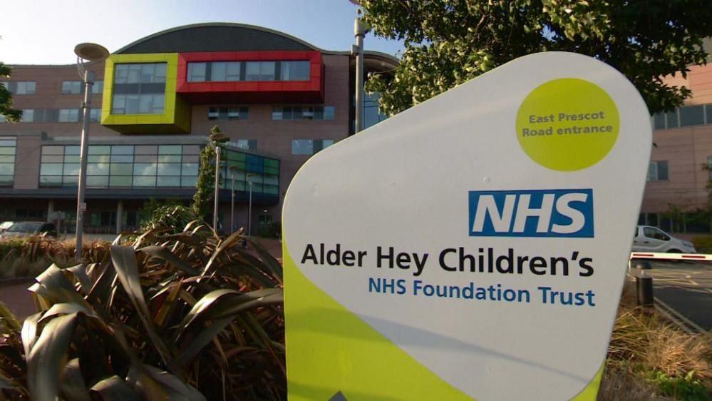 A sign outside the front of a large brick and glass building reads 'Alder Hey Children's NHS Foundation Trust'. 
