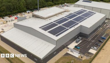 An aerial view of a large grey and black facility with car parks on both sides of the building.