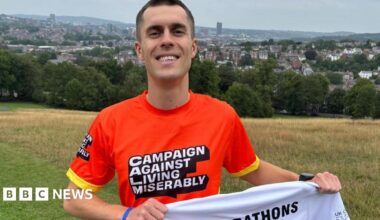 A smiley man in a bright orange sports shirt stands on a grass field with the city spread out behind him