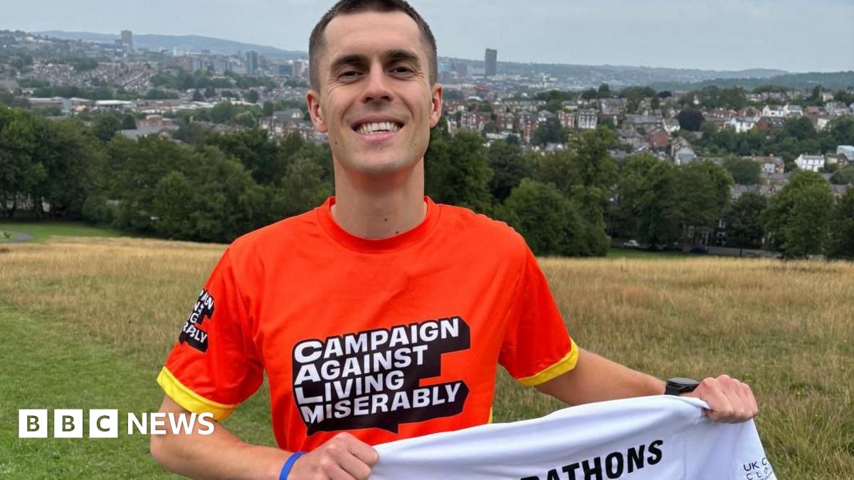 A smiley man in a bright orange sports shirt stands on a grass field with the city spread out behind him