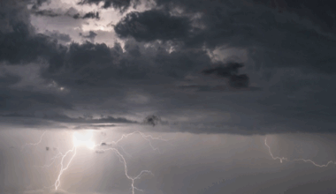 Shows grey skies with bolts of lightning above water and lights and mountains in the background