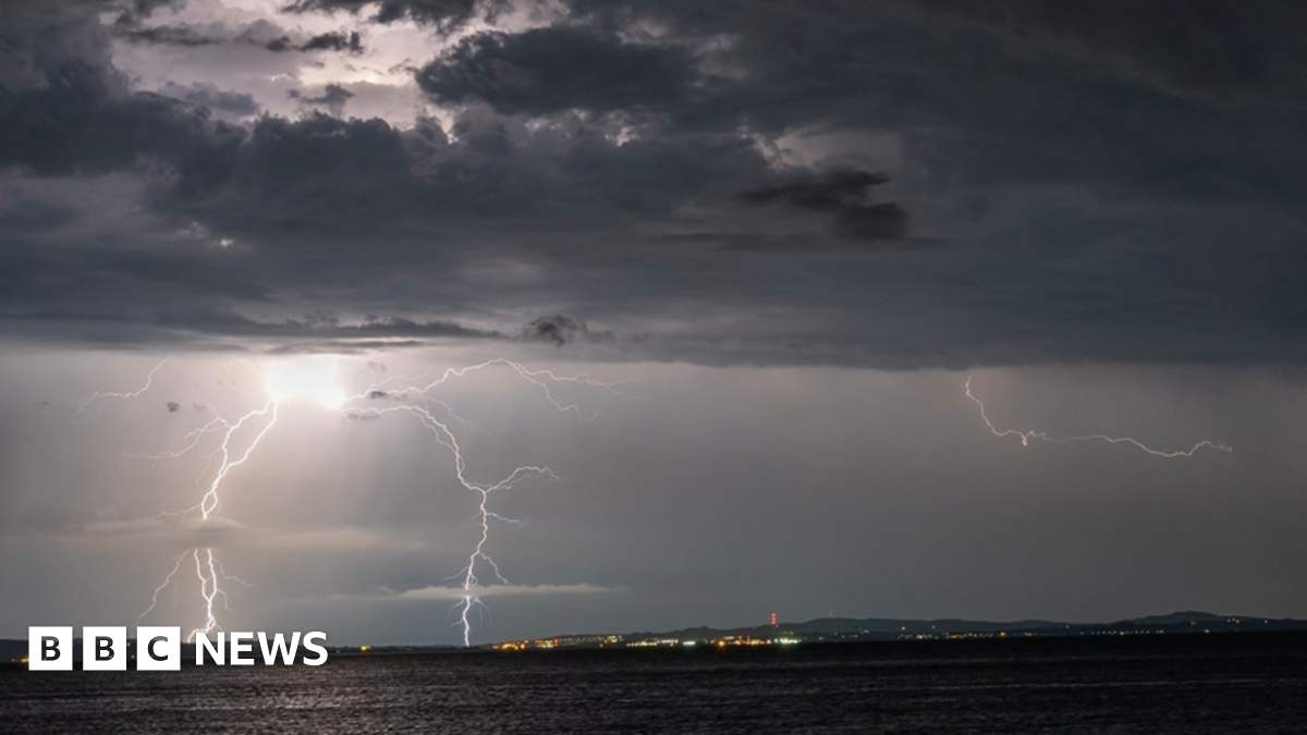Shows grey skies with bolts of lightning above water and lights and mountains in the background