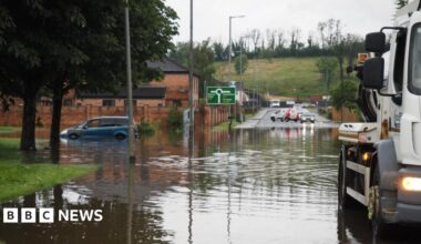 Flooding on the main Enniskillen to Dublin Road in Enniskillen on Sunday evening.  Part of the road is submerged in flood water. One blue hatchback car is parked on the left of the photo.  The water level is up to its rear bumper and more than half way up its rear wheel arch. A large white truck used to clear drains is in the foreground and cars are turning away from the area in the background.
