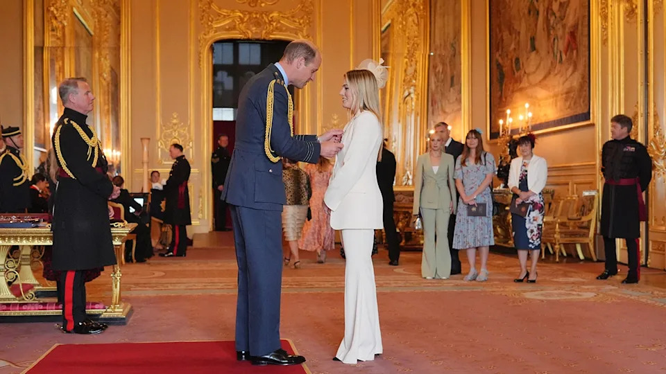 Prince William in a blue suit pinning a medal to a woman who is wearing a white suit.