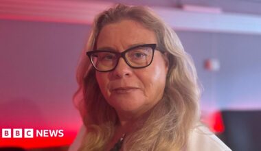 A head and shoulders shot of Marcella Leonard. She has longer, blonde hair and is wearing black-framed cat eye glasses. She is wearing a white blazer and a black shirt. She is looking down the camera lens with a neutral expression on her face.