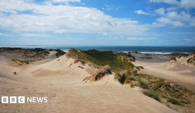 Sand dunes with a couple of people walking in the distance and the blue sea and sky in the background.