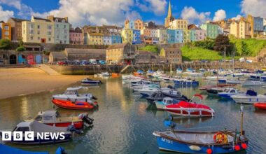 A wide shot of Tenby harbour, with colourful buildings nestled in the cliff overlooking the water. A number of boats are lined up at the front of the shot with the beach on the left hand side.