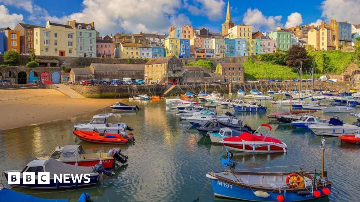 A wide shot of Tenby harbour, with colourful buildings nestled in the cliff overlooking the water. A number of boats are lined up at the front of the shot with the beach on the left hand side.