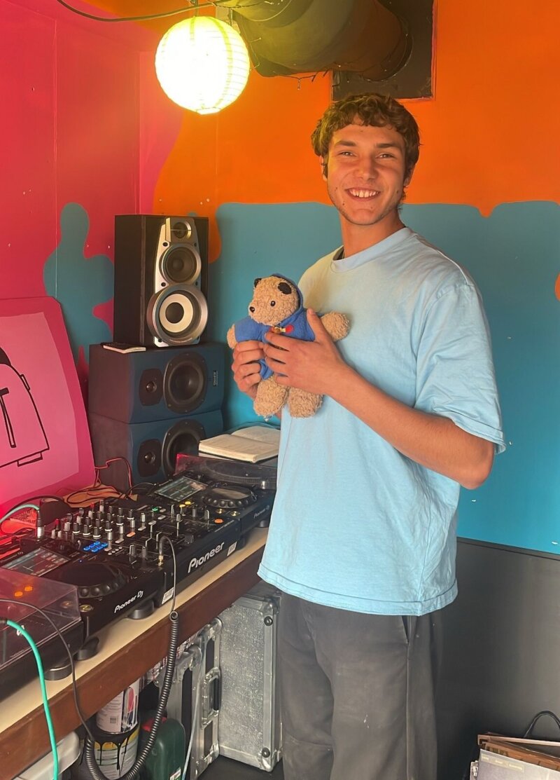 A photo of a smiling man in a baby blue t-shirt holding a teddy bear, standing next to some DJ equipment and decks in a brightly painted room