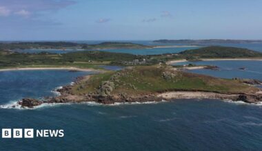 The island of Bryher is shown with rocky hills, sandy beaches and bodies of water beneath a blue sky scattered with clouds.