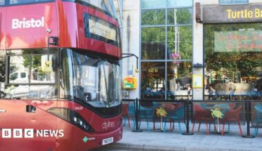 A red bus with "Bristol" on the side and "First Bus" on the front sits in front of a restaurant called Turtle Bay. The restaurant has seating on the pavement.