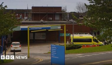 The entrance to Furness General Hospital. A blue sign shows directions to different departments in front of a road heading down to the main entrance, above which is a sign containing the hospital's name. A white car and an ambulance are parked outside. Scaffolding sits on an upper part of the low, brick-built building.