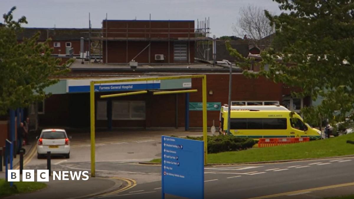 The entrance to Furness General Hospital. A blue sign shows directions to different departments in front of a road heading down to the main entrance, above which is a sign containing the hospital's name. A white car and an ambulance are parked outside. Scaffolding sits on an upper part of the low, brick-built building.