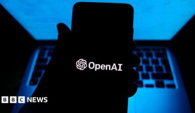A person holding a phone with OpenAI software in a dark room with a computer behind their hand