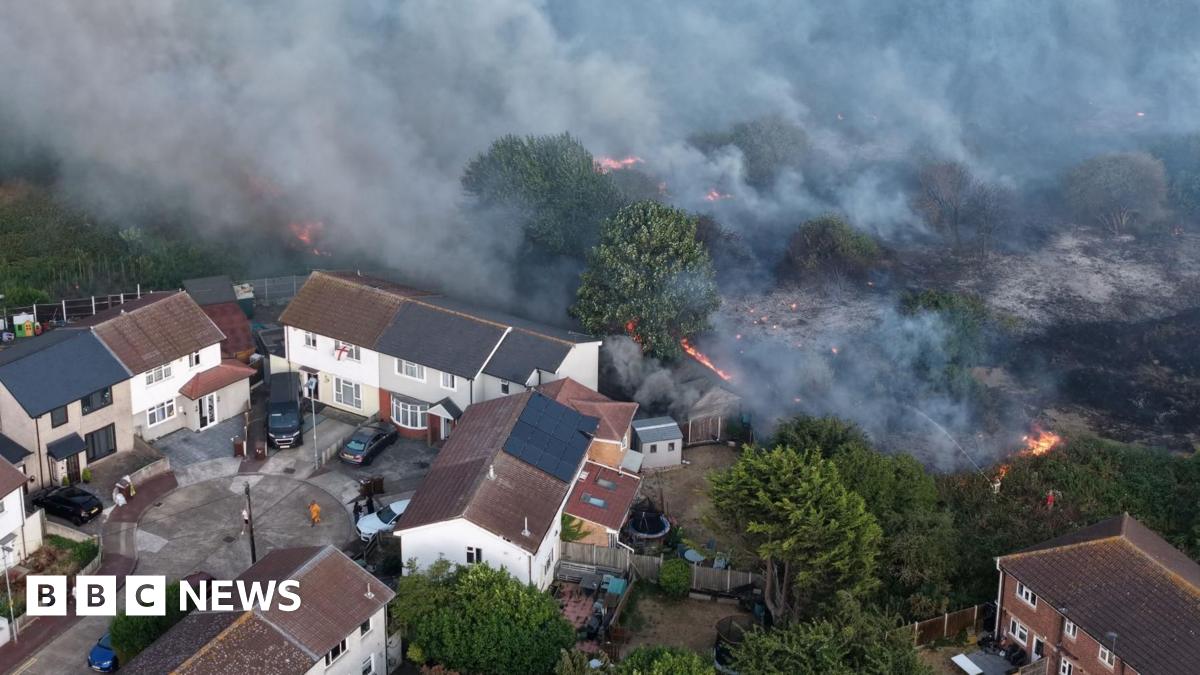 A fire burning on grass near trees and a cluster of houses
