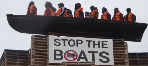 PA Media The picture shows a model of a small boat with figures inside in orange fluorescent vests. Below is a sign which says 'STOP THE BOATS'.