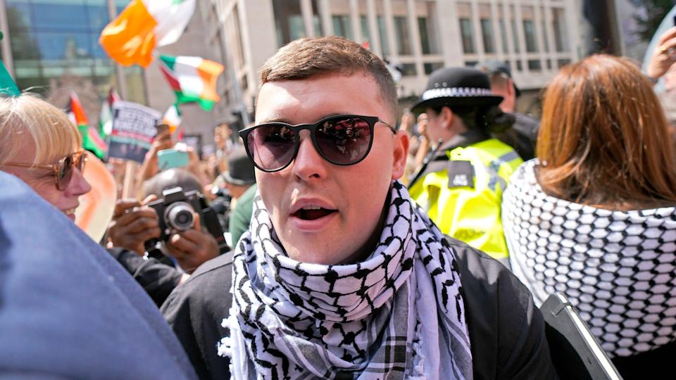 A man wearing glasses and a keffiyeh, stands outside court - in the background dozens of supporters, some waving Irish flags, can be seen