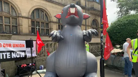 BBC A giant inflatable grey rat is tethered in front of Sheffield Town Hall. In the background are trade union banners and flags.