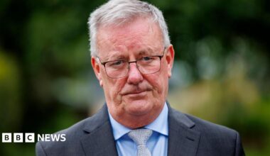 Mike Nesbitt, he has grey hair and is wearing glasses and a grey suit jacket over a blue shirt and silver tie.