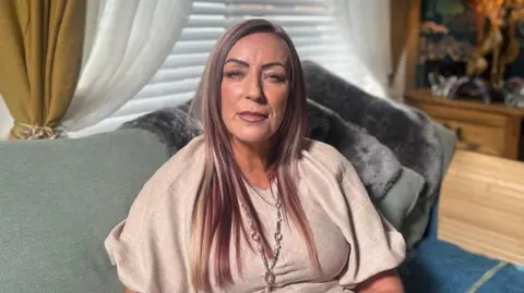 BBC Julieanne Boyle sitting on a green sofa in a living room looking directly at the camera.  She has long, straight hair with blonde and copper tones. She is wearing a loose beige top, and a long silver and pearl necklace. 