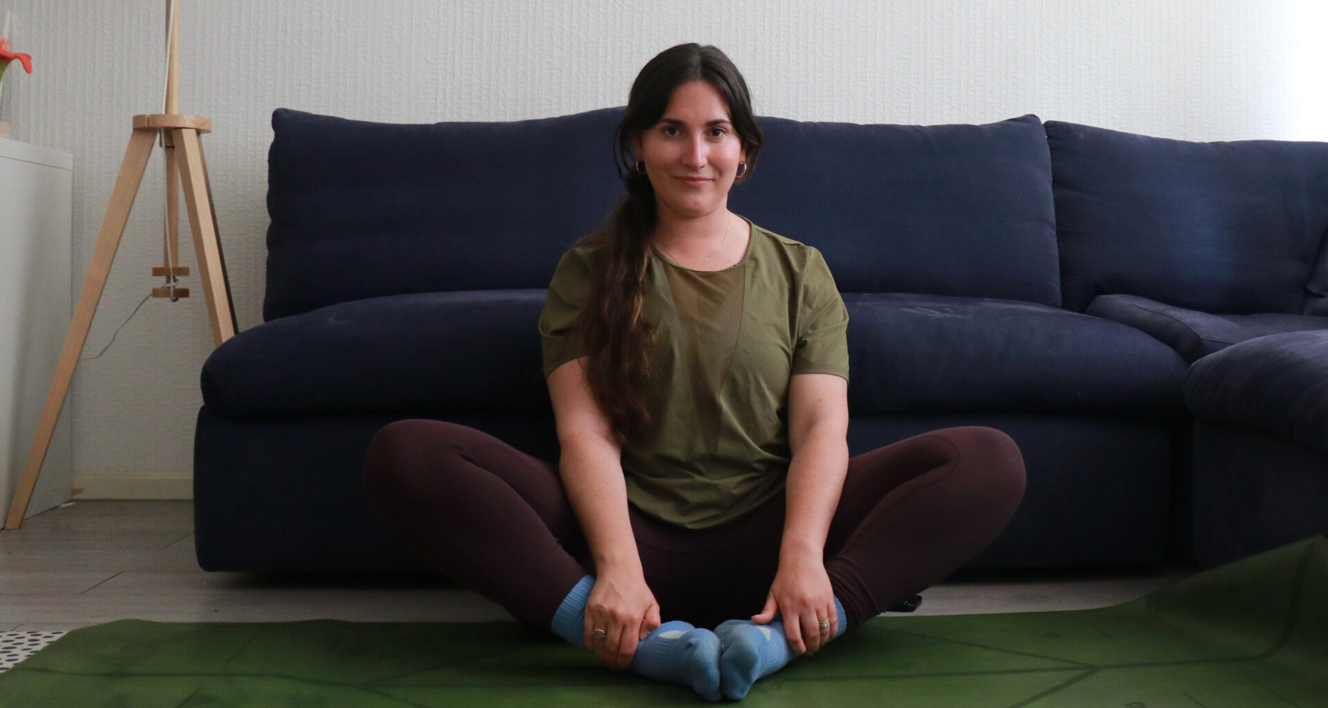 Marina Leiva García performs a butterfly pose at home. She is sitting with her knees bent and the soles of her feet together. Her knees are pointing upwards at a 45° angle. Behind her is a couch.