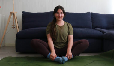 Marina Leiva García performs a butterfly pose at home. She is sitting with her knees bent and the soles of her feet together. Her knees are pointing upwards at a 45° angle. Behind her is a couch.