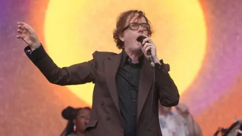 PA Media Jarvis Cocker, wearing glasses and a brown jacket with Pulp performing on the Pyramid Stage during the Glastonbury Festival at Worthy Farm in Somerset. 