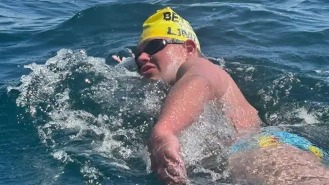 Oscar is in water, he is wearing a yellow swim cap, black goggle and blue and yellow swim pants. He is breathing to his left side as he takes a stroke. 
