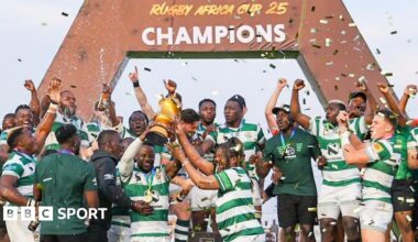 Zimbabwe's rugby players dressed in green and white lift a golden trophy and celebrate in front of a sign that says Rugby Africa Cup 25 champions