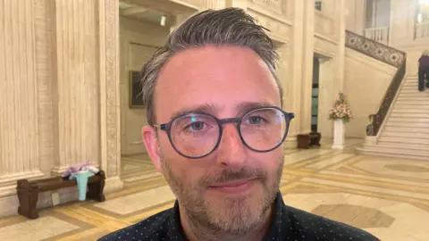 Nick Matheson has a grey beard, grey/whitish hair that is gelled into a spike and grey round glasses. He is wearing a navy shirt and behind him is the yellow white panelled hall and stairs of Stormont. 