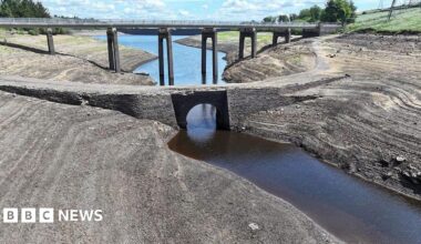 A dried-up bed of a reservoir with only a narrow stream of water flowing through the middle. In the background there is a bridge and a blue sky with scattered clouds.