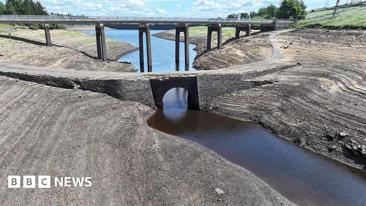 A dried-up bed of a reservoir with only a narrow stream of water flowing through the middle. In the background there is a bridge and a blue sky with scattered clouds.