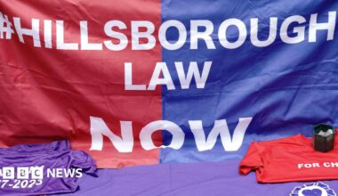 A Hillsborough Law banner. It reads #Hillsborough Law Now - blue on one half and red on the other with white lettering. Two t-shirts - one purple, the other red - are in front of it, along with a candle.