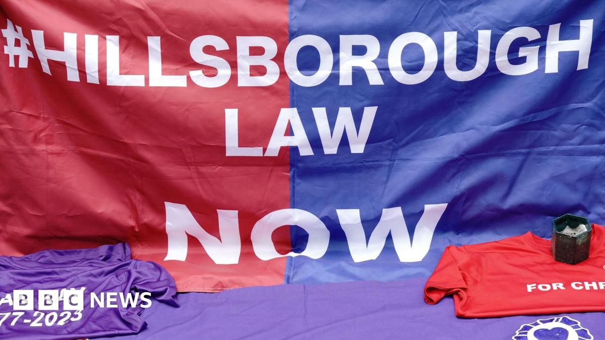 A Hillsborough Law banner. It reads #Hillsborough Law Now - blue on one half and red on the other with white lettering. Two t-shirts - one purple, the other red - are in front of it, along with a candle.
