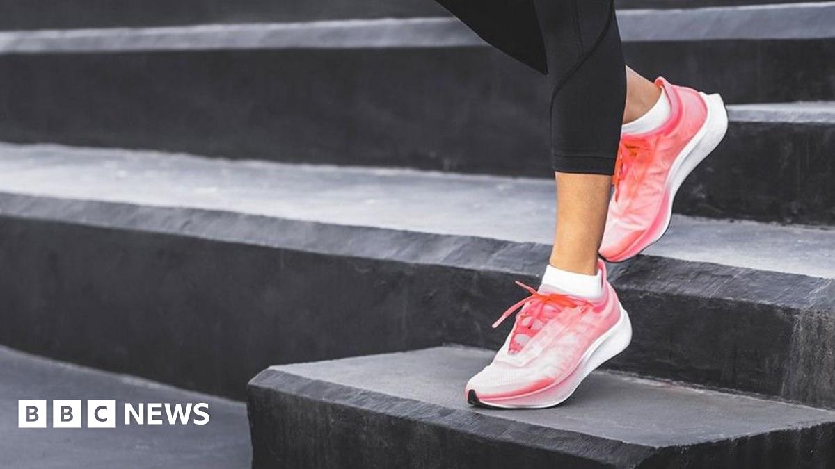 A woman wearing black leggings and pink athletic shoes, standing on a set of black steps. One foot is placed on a higher step while the other remains on a lower one, suggesting an exercise or stretching.