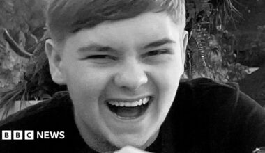 Harvey Willgoose laughing at the camera with his mouth open, wearing a black top. The photo is in black and white.