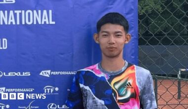 A young man with short dark hair and a multi-coloured sports top standing in front of a blue advertising screen with a clay tennis court partially visible behind him